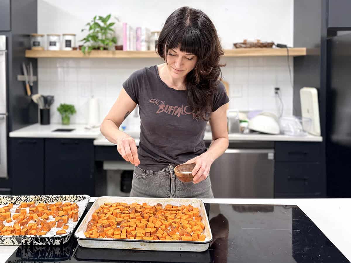 Una mujer espolvoreando sal sobre una bandeja de batatas cortadas en cubitos en una cocina.