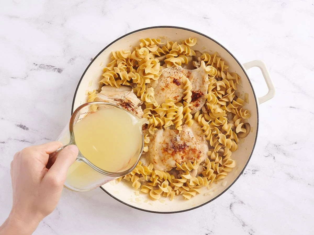 Fideos en una sartén con muslos de pollo y champiñones, y vierte el caldo.