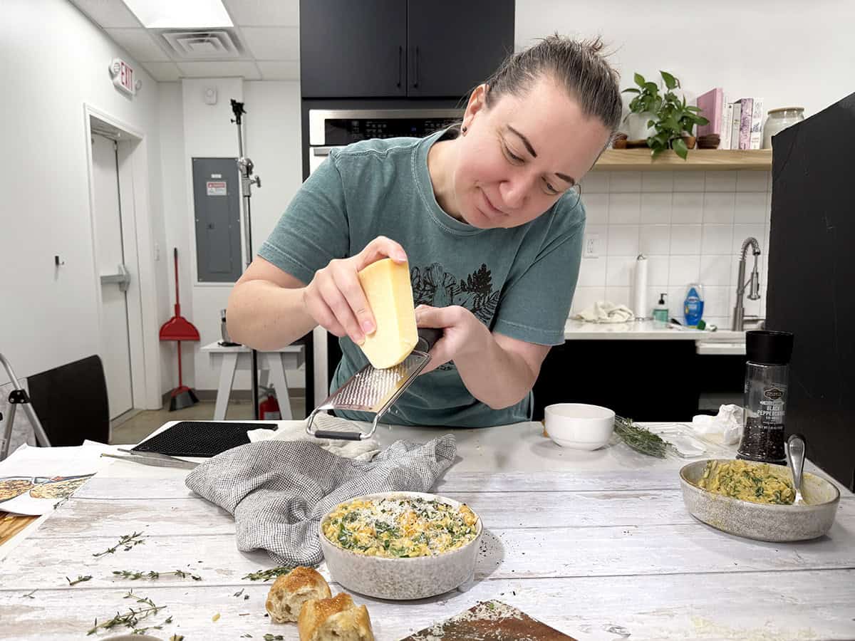 Una mujer triturando parmesano fresco sobre un recipiente sobre orzo de calabaza en una cocina.