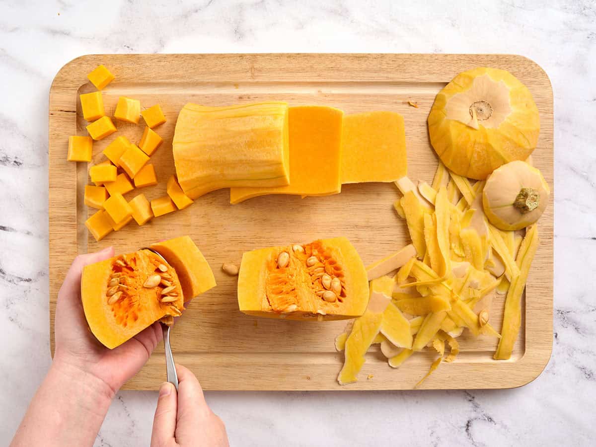 Manos preparando calabaza quitando las semillas, rodeadas de cáscara de calabaza y calabaza cortada en cubitos sobre una tabla de cortar.